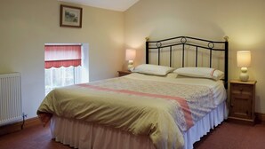Un dormitorio con una cama, dos mesitas de noche, una ventana con cortinilla roja, un radiador y un cuadro en la pared.