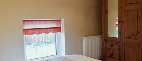 A bedroom with a bed, a window with a red valance, a radiator, and a wooden wardrobe.