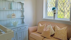 Ein Zimmer mit einem hellblauen Schminktisch, einem beigen Sofa mit gemusterten Kissen und einem Fenster mit Gitterwerk, sowie einer Vase mit Blumen.