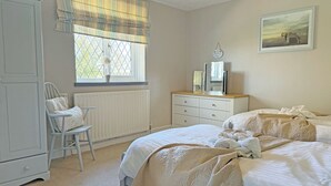 Ein Schlafzimmer mit Bett, Kommode, Stuhl, Fenster mit Vorhängen, Heizkörper und einem gerahmten Bild an der Wand.