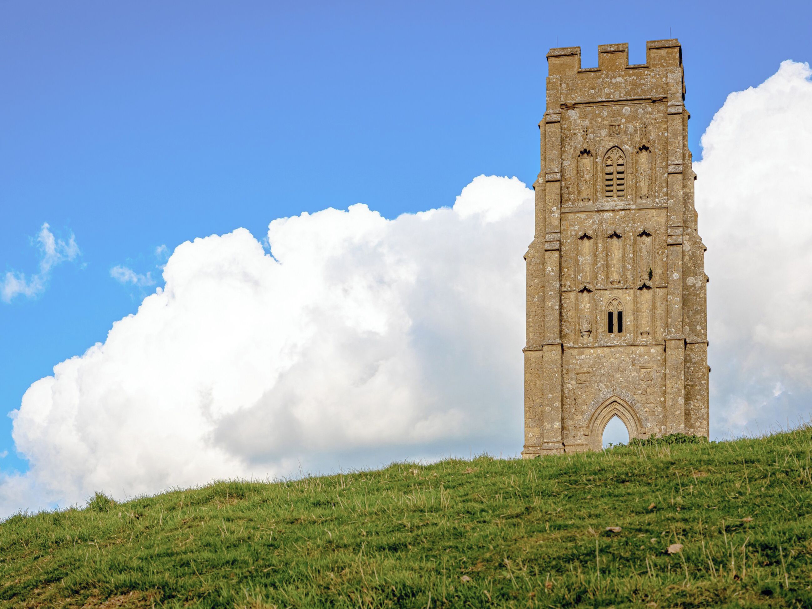Ein historischer Stein-Turm auf einem grasbewachsenen Hügel, bewölkter Himmel.