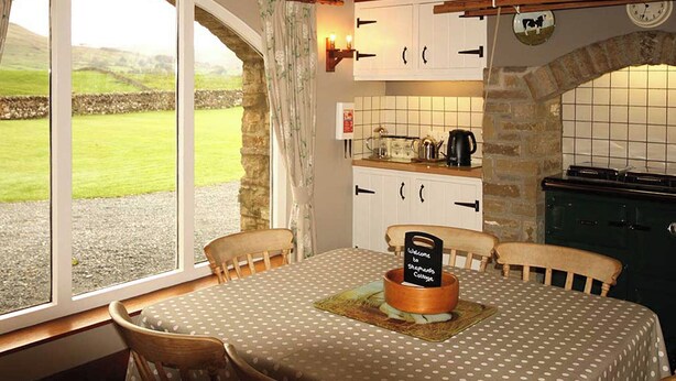 Interior acogedor de una cabaña con una mesa de comedor, sillas, una zona de cocina y vistas al campo a través de un ventanal grande.