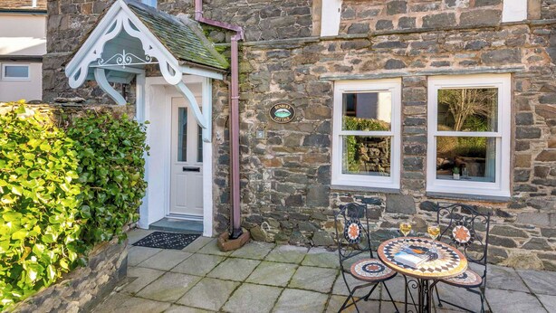 A stone building with a white door, a sign reading 'Ribnure Cottage,' and a small outdoor seating area with a table and chairs. - 1 bedroom accommodation in Keswick (Keswick)
