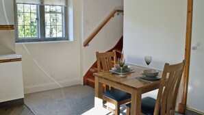 Un comedor con una mesa de madera preparada para dos personas, una ventana con persianas, una escalera y una zona de cocina con encimera blanca.