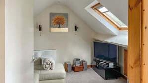 Un dormitorio estilo loft acogedor, con techo inclinado, un ventanal grande, un televisor de pantalla plana, un sofá blanco y un cuadro de un árbol.