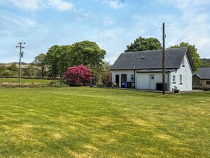 A white house with a dark roof, surrounded by a grassy field and a large tree with pink flowers.