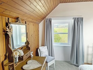 Ett rum med ett träpanelat tak, ett stort fönster med gardiner, en spegel, en byrå i trä, en stol och ett litet bord med en vas med blommor.