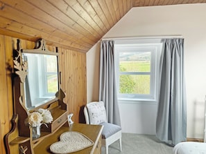 Une pièce avec un plafond en bois, une grande fenêtre avec des rideaux, un miroir, une commode en bois, une chaise et une petite table sur laquelle est posé un vase de fleurs.
