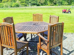 Une table et des chaises en bois sont disposées à l’extérieur, avec un champ verdoyant et un tracteur en arrière-plan.