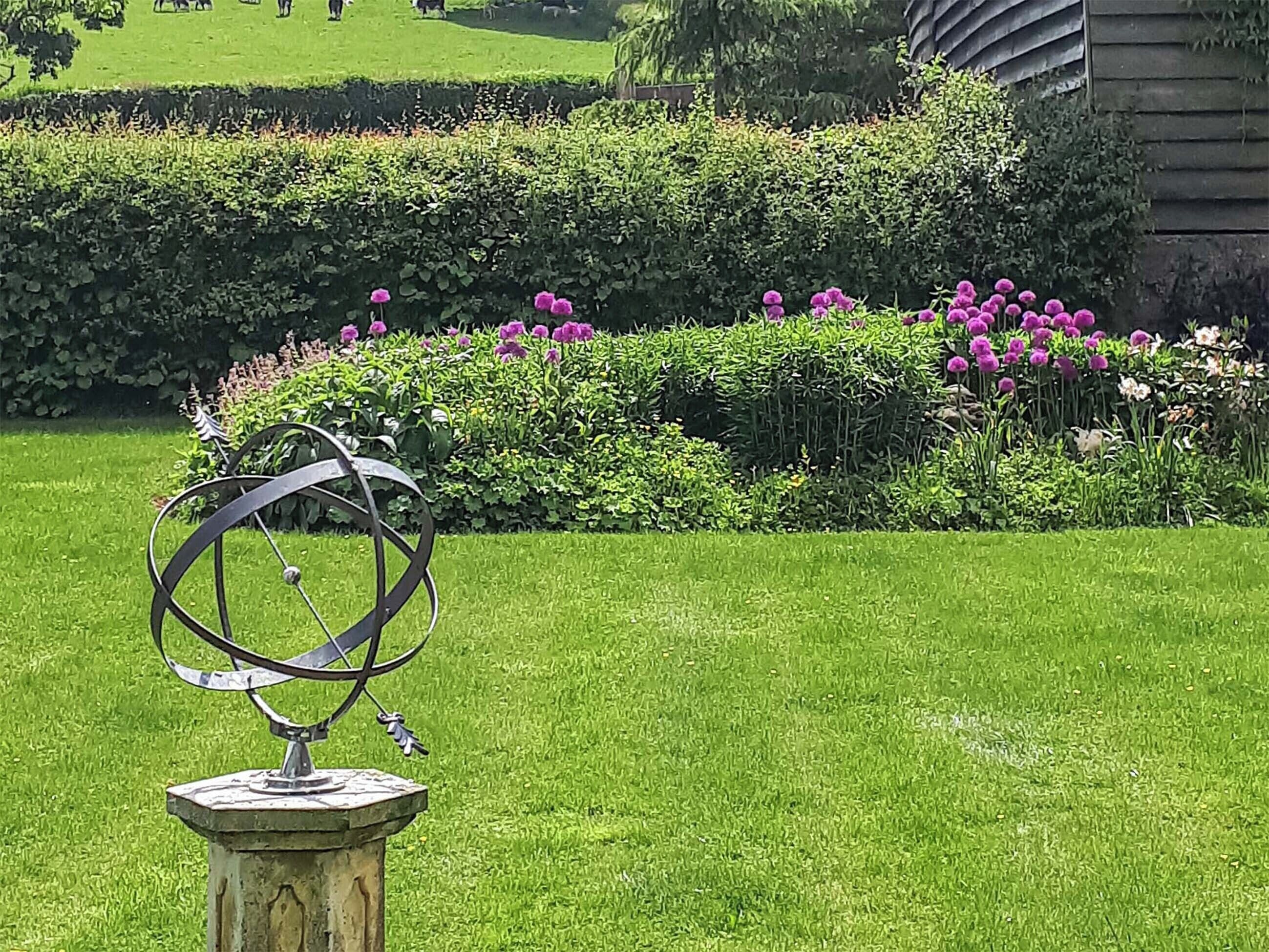 Un jardin agrémenté d’une sculpture métallique posée sur un piédestal, entouré de pelouse bien entretenue et d’arbustes en fleurs.