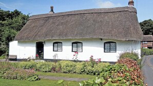 Ein weißes Cottage mit Strohdach, einer Holztür und einem kleinen Garten mit Blumen und Sträuchern.