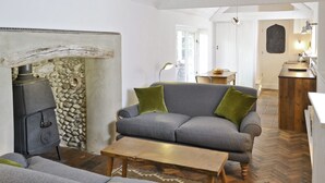 Una sala de estar acogedora con una chimenea de piedra, un sofá gris con cojines verdes, una mesa de centro de madera y un suelo con estampado de espiga.