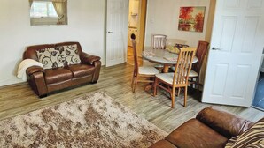 A living room with a brown leather sofa, a dining table with chairs, a patterned rug, and a painting on the wall. - 1 bedroom accommodation in Horning, near Norwich (Horning, near Norwich)
