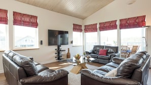A living room with leather sofas, a flat-screen TV, a coffee table, and large windows with red curtains. - Swedish timber-style house, with sauna and hot tub, on a 400-acre estate. (Fritton, near Great Yarmouth)