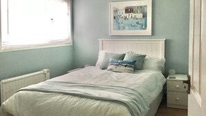 A bedroom with a bed, a nightstand, a bedside table, a framed picture on the wall, and a window with blinds. - Enjoy distant sea views whilst relaxing amidst the stunning rural surroundings. (St Merryn, near Padstow)