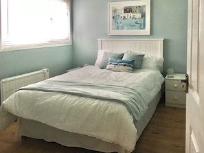 A bedroom with a bed, a nightstand, a bedside table, a framed picture on the wall, and a window with blinds. - Enjoy distant sea views whilst relaxing amidst the stunning rural surroundings. (St Merryn, near Padstow)
