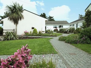 A white house with a paved driveway leading to a front door, surrounded by a well-maintained garden with various plants and flowers. - Enjoy distant sea views whilst relaxing amidst the stunning rural surroundings. (St Merryn, near Padstow)