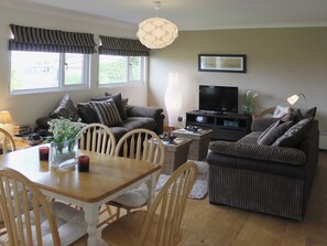 A living room with a sofa, a TV, a dining table with chairs, and a window with curtains. - Enjoy distant sea views whilst relaxing amidst the stunning rural surroundings. (St Merryn, near Padstow)