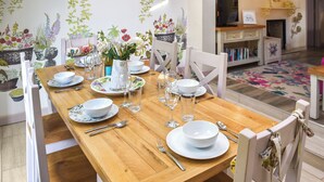 A dining room with a wooden table set for four, white chairs, and a television in the background. - Captain's Cottage - 27888 (Winterton-on-Sea)