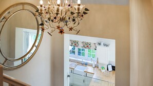 A staircase leading to a kitchen with a chandelier, a large mirror, and a window with floral curtains. - Captain's Cottage - 27888 (Winterton-on-Sea)