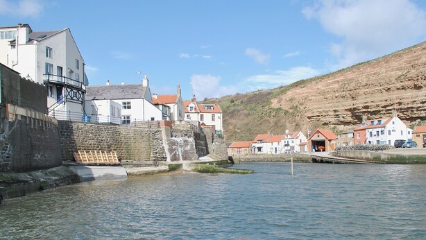 A waterfront town with stone walls, a small waterfall, and traditional buildings with red roofs. - The Little Stone Cottage (Loftus, near Saltburn-by-the-Sea)