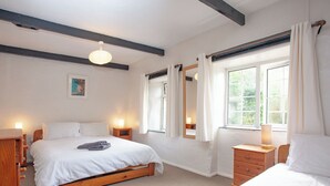 A bedroom with two beds, a wooden dresser, a nightstand with a lamp, a round rug, and a window with curtains. - 1 bedroom accommodation in Looe (Looe)