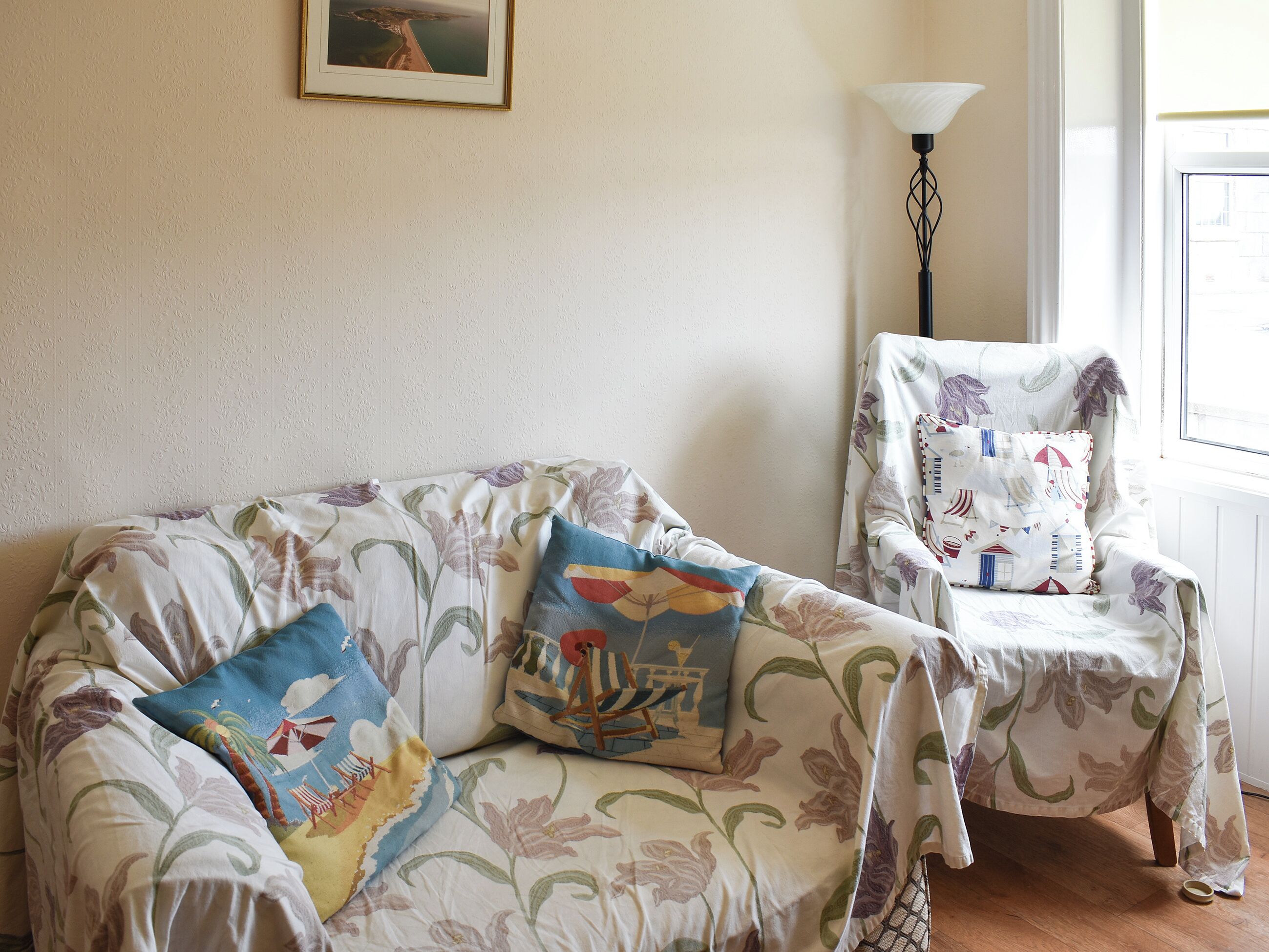 A living room with floral-patterned furniture, a framed picture on the wall, and a lamp near a window.
