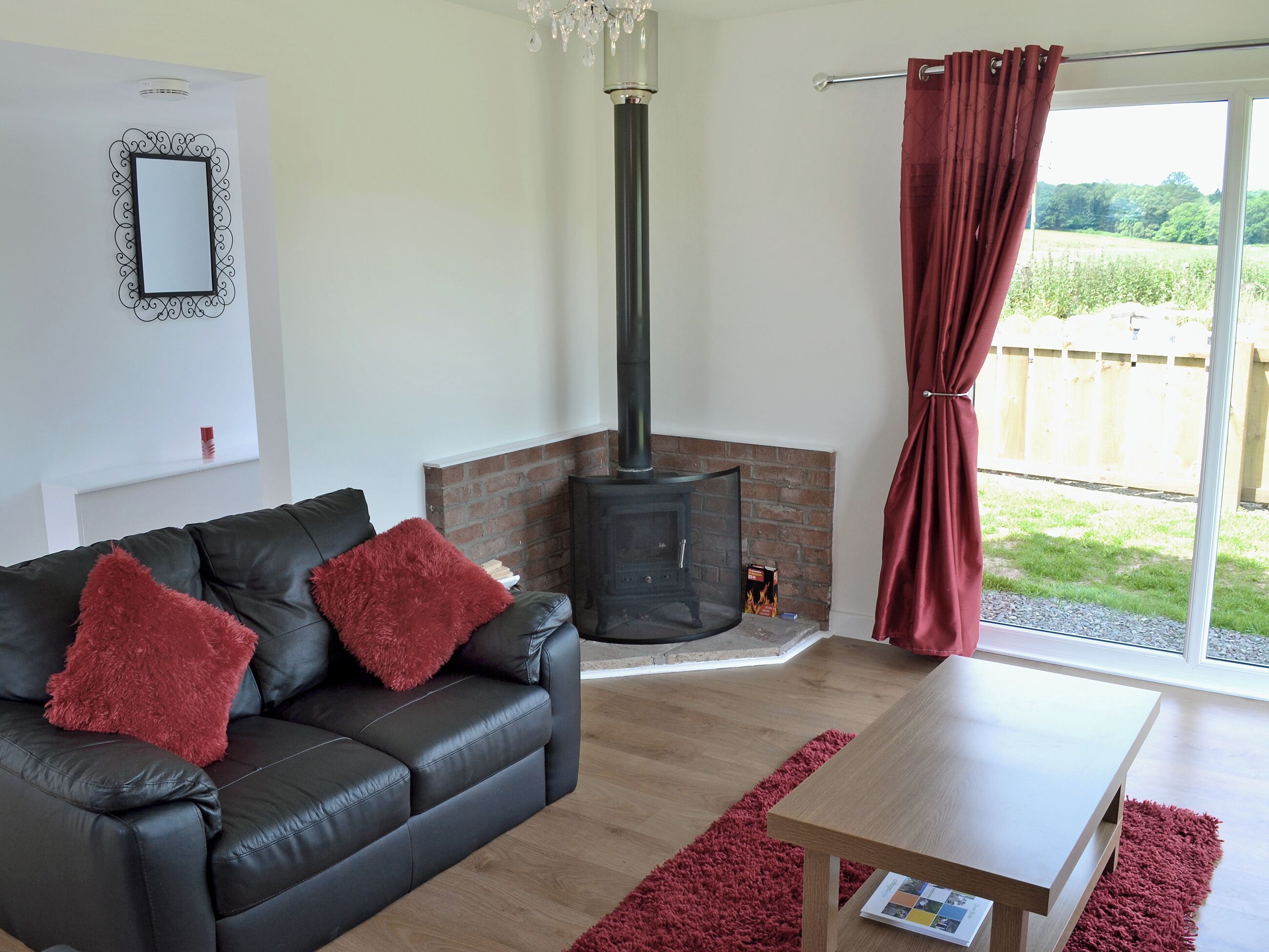 Ein Wohnzimmer mit einer schwarzen Ledersofa, roten Kissen, einem Holztisch, einem Glastischkamin und einem großen Fenster mit Blick ins Grün.