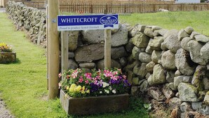 Een bordje voor Whitecroft Scotch Assured Farm, omringd door een stenen muur en kleurrijke bloemen.