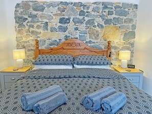 Una cama con ropa de cama estampada, dos lámparas de noche, un reloj digital y una pared de piedra al fondo.