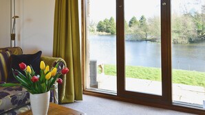 Una habitación con vistas a un lago, una mesa con un jarrón de tulipanes, un sofá con un cojín decorativo y una lámpara de pie.