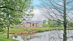 Una casa junto a un lago con un puente de piedra, rodeada de hierba y árboles.