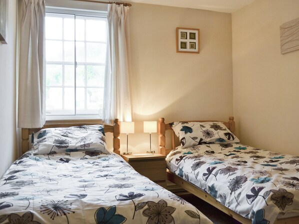 A room with two single beds, a window with curtains, a bedside table with a lamp, and a small wall-mounted picture.