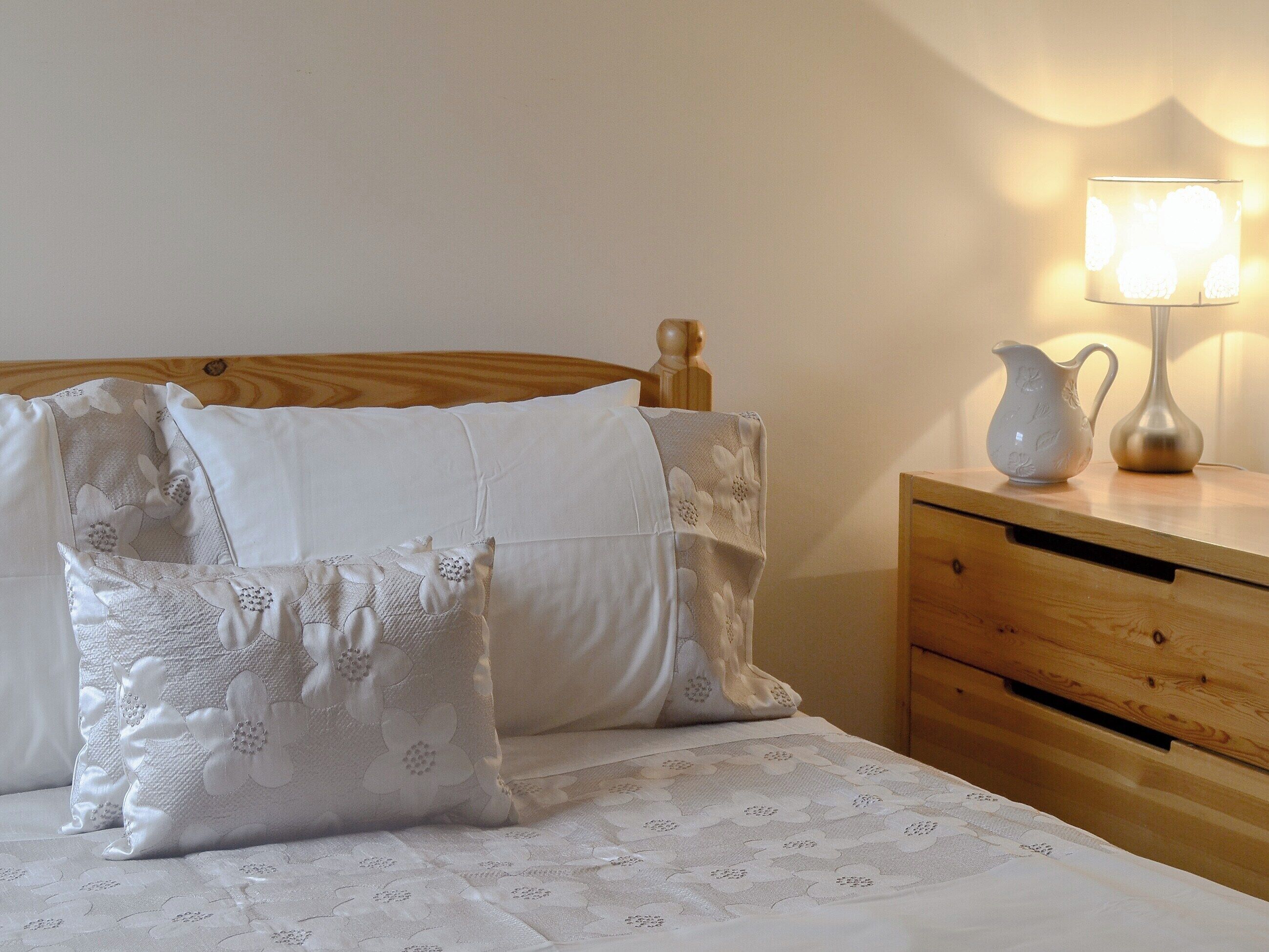 Un lit bien fait, avec du linge de lit blanc et des coussins à motifs floraux, une table de chevet en bois avec une lampe, et une tête de lit en bois.