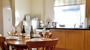 A dining area with a wooden table set for four, surrounded by wooden chairs. A kitchen is visible in the background with wooden cabinets and a window with blinds.