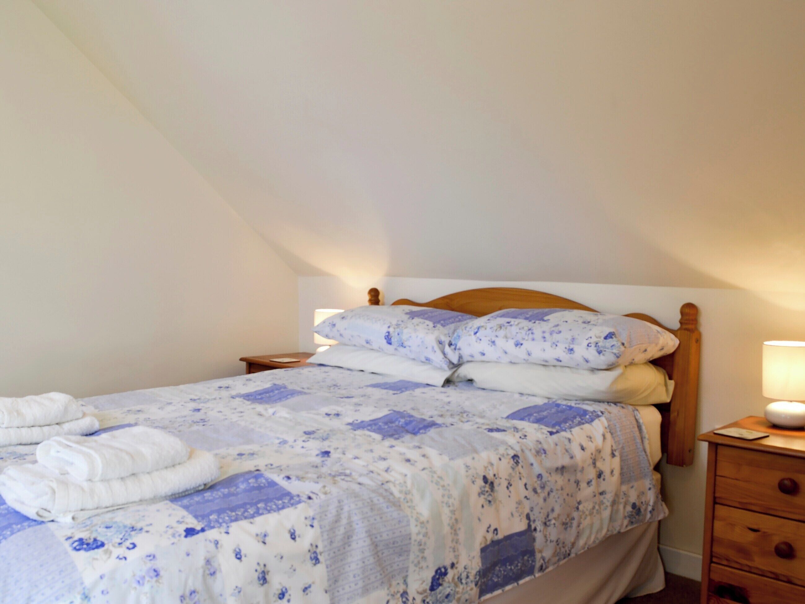 A neatly made bed with a floral bedspread and pillows, a wooden headboard, a bedside lamp, and a wooden nightstand with a drawer.