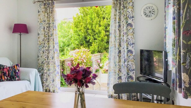 A room with a wooden table, a TV on a stand, a bed with a colorful pillow, and a window with curtains. - 2 bedroom accommodation in Bothenhampton, near Bridport (Bothenhampton, near Bridport)