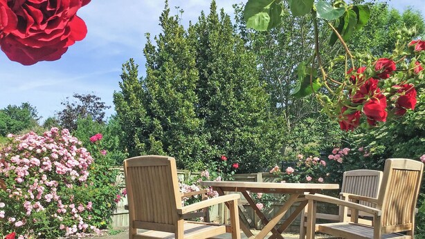 Un espacio de estar al aire libre con madera, que incluye una mesa y sillas, rodeado de abundante follaje y plantas en flor.