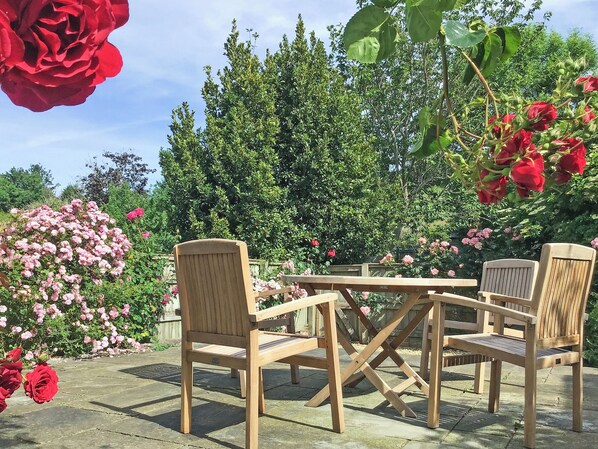 A wooden outdoor seating area with a table and chairs, surrounded by lush greenery and flowering plants.