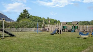 A playground with slides and climbing structures, surrounded by a wooden fence and greenery.