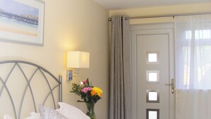 A hotel room with a bed, a wall-mounted lamp, a vase with flowers, a door with a window, and a framed picture on the wall.