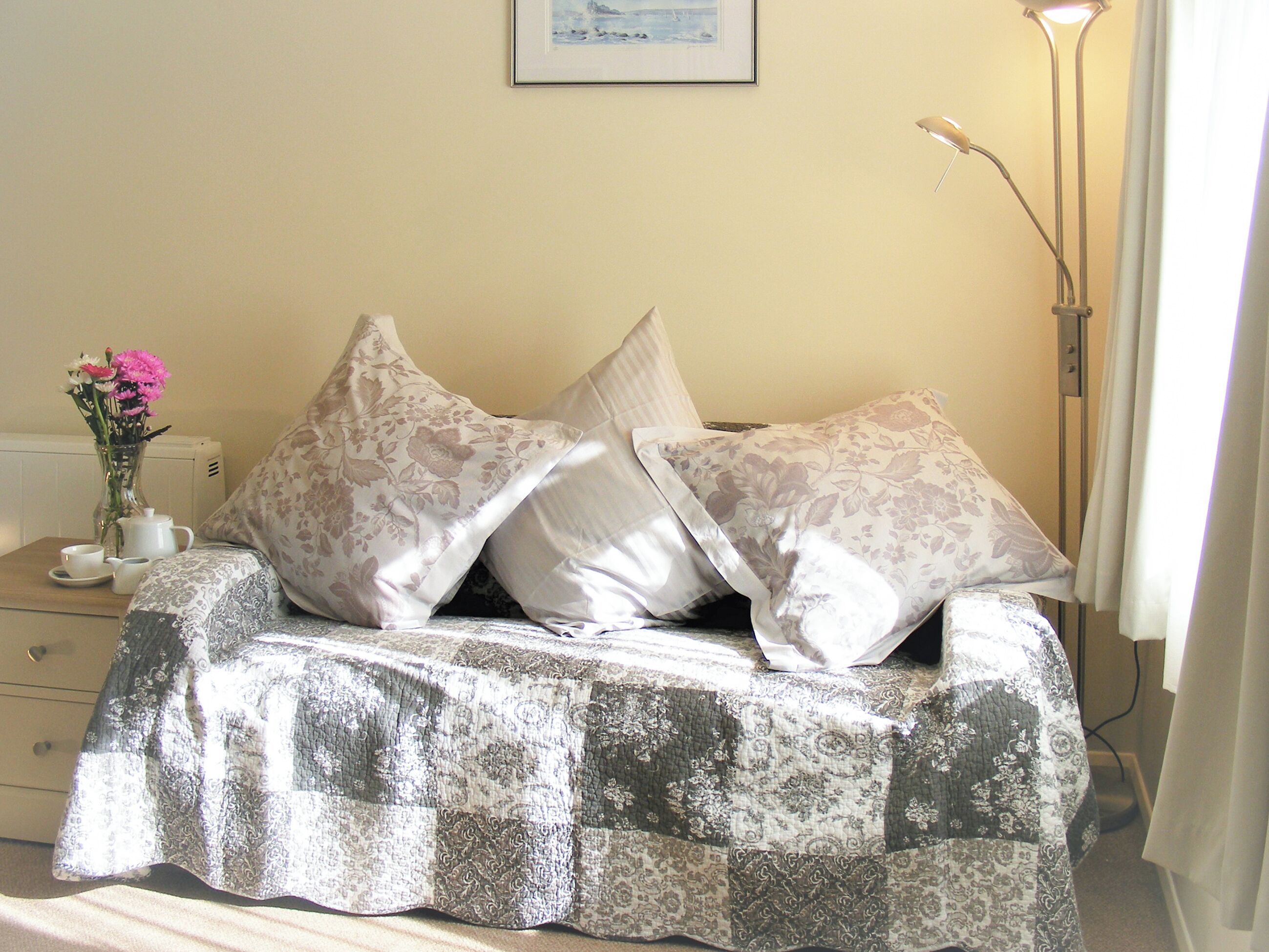 A bed with a patterned bedspread, a bedside table with a vase of flowers, and a floor lamp.