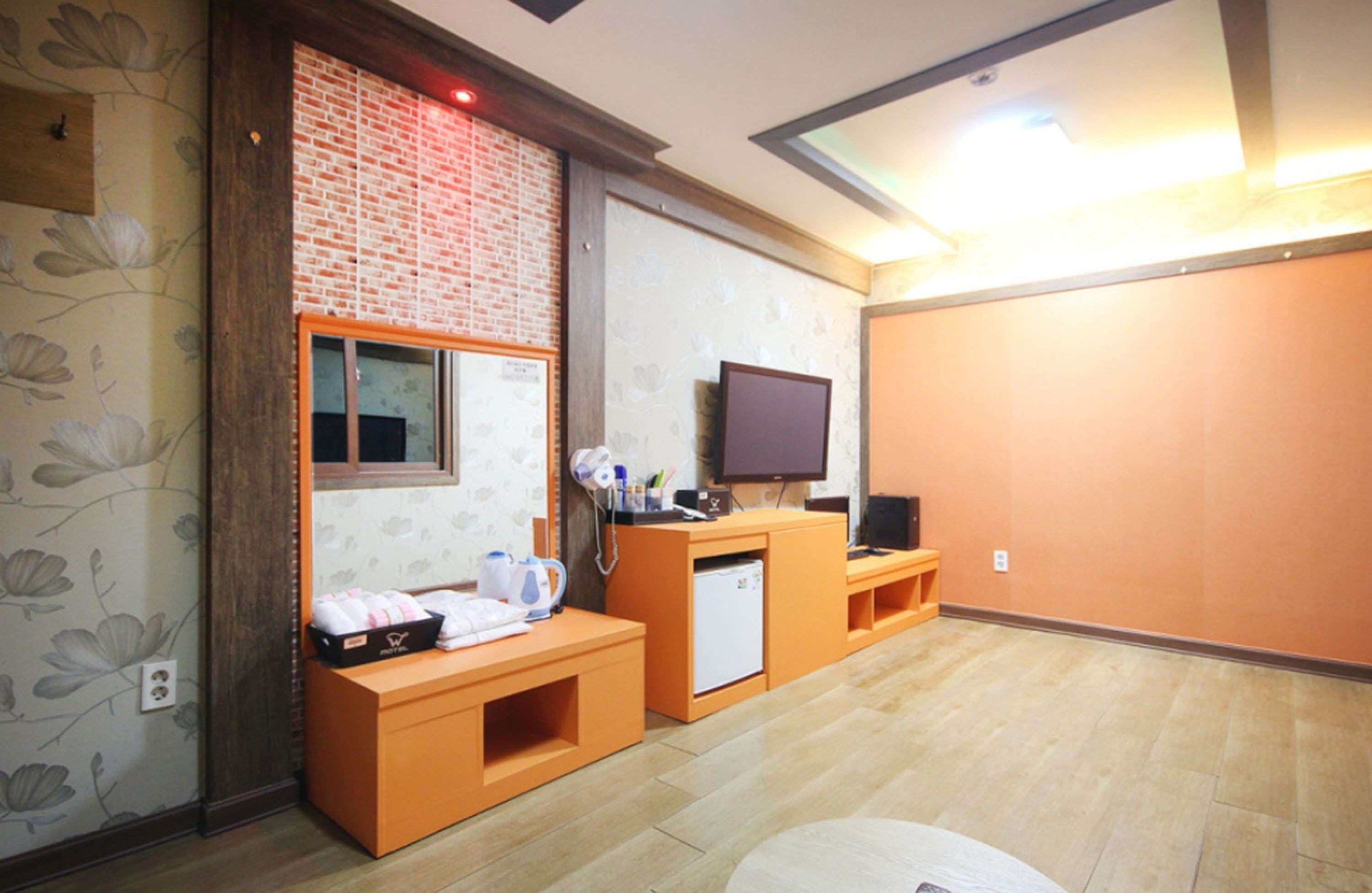 A hotel room with a wooden floor, a brick accent wall, a television, a small refrigerator, and a desk with a kettle and cups.