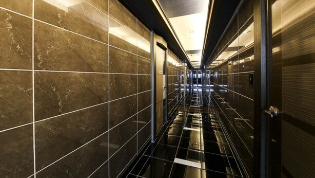 A modern, well-lit hallway with reflective black and gold tiles, leading to a glass elevator. - W Motel (Tongyeong)