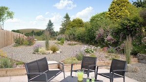 Un patio al aire libre con tres sillas y una mesa, rodeado de un jardín con diversas plantas y flores.