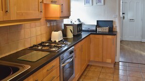A kitchen with wooden cabinets, a gas stove, a toaster, and a microwave. There is a tiled floor and a white door in the background. - 3 bedroom accommodation in Bala (Bala)