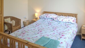A bedroom with a wooden bed, a wicker chair, a bedside table with a lamp, and a wall socket. - 1 bedroom accommodation in Nancledra, near Penzance (Nancledra, near Penzance)