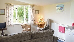 A room with a sofa, a chair, a desk, a radiator, and a window with curtains. - 1 bedroom accommodation in Nancledra, near Penzance (Nancledra, near Penzance)