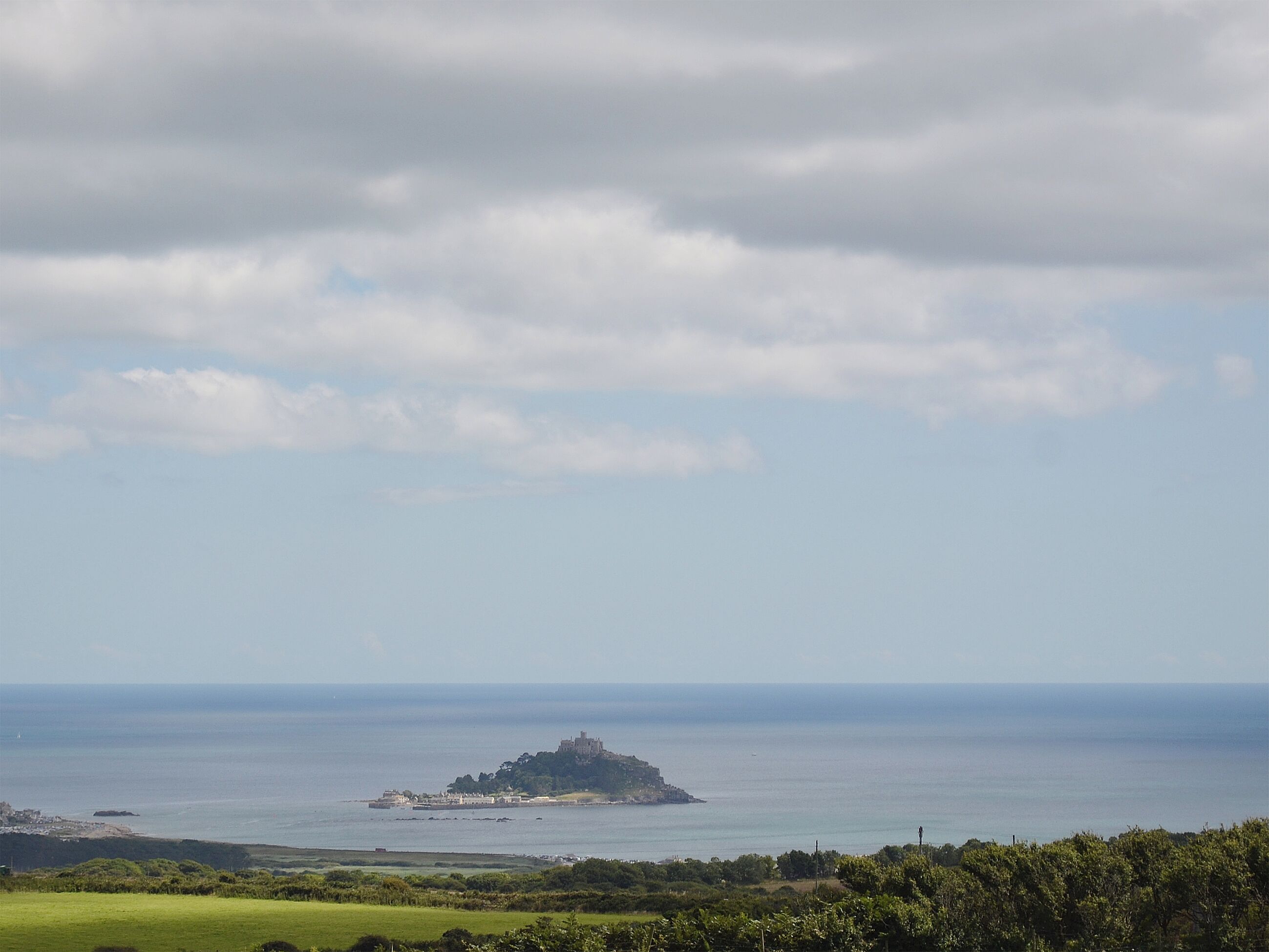 Eine Küstenlandschaft mit einer Burg auf einer kleinen Insel, einer entfernten Stadt und wolkenlosem Himmel.