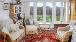 A cozy room with wicker furniture, a red patterned rug, and large windows offering a view of a green field. - 1 bedroom accommodation in Stalham (Stalham)